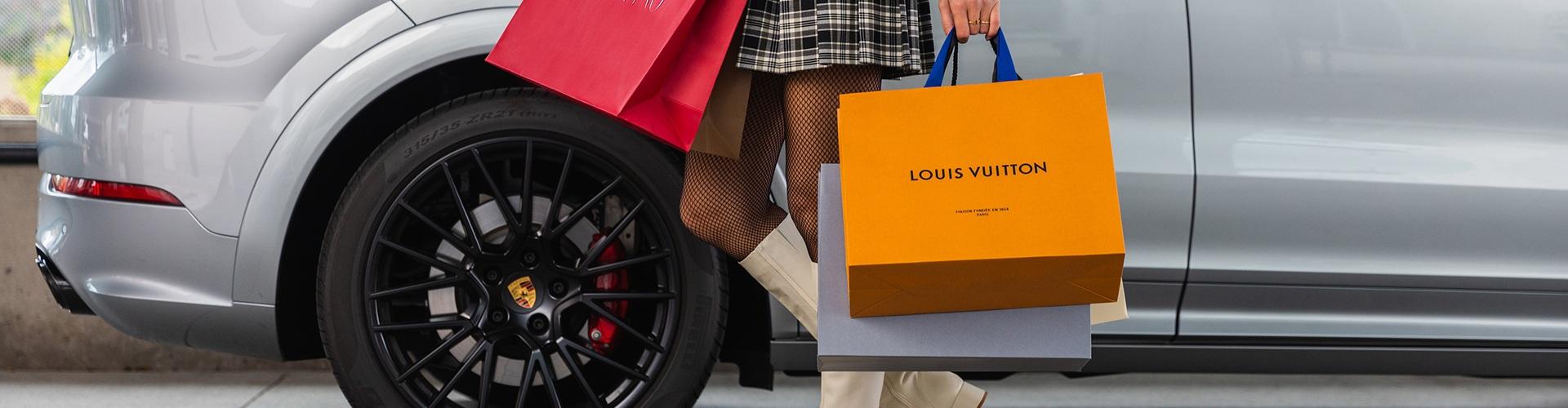 Woman in plaid skirt carrying Louis Vuitton shopping bags beside a Porsche