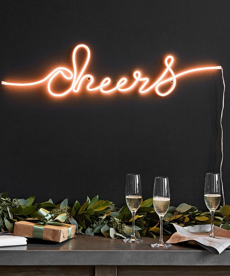 A hanging neon light from Pottery Barn that says "cheers". Below it is a tabletop with greenery, gifts, and three glasses of champagne.