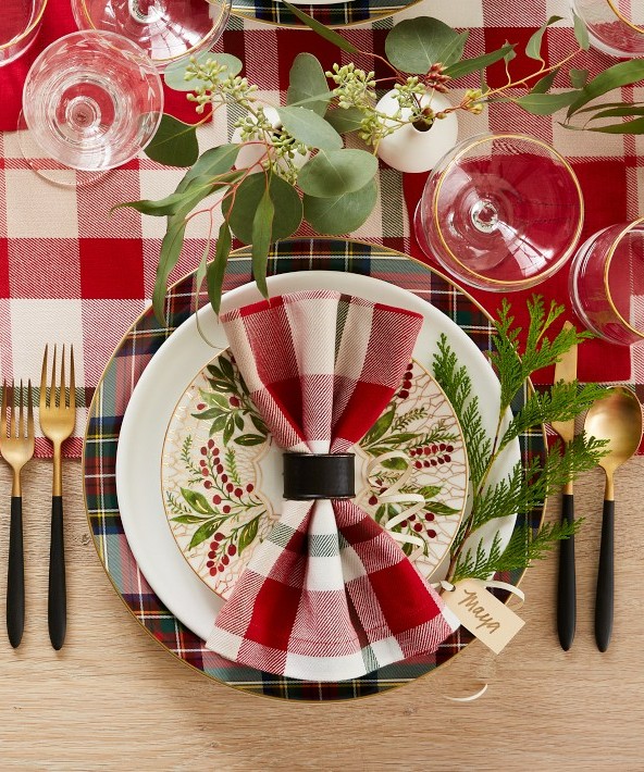 A white William Sonoma dinner plate with red and green tartan pattern on the rim, set on top of a larger tartan charger plate. A checkered napkin is folded and placed nicely on top of the plates.