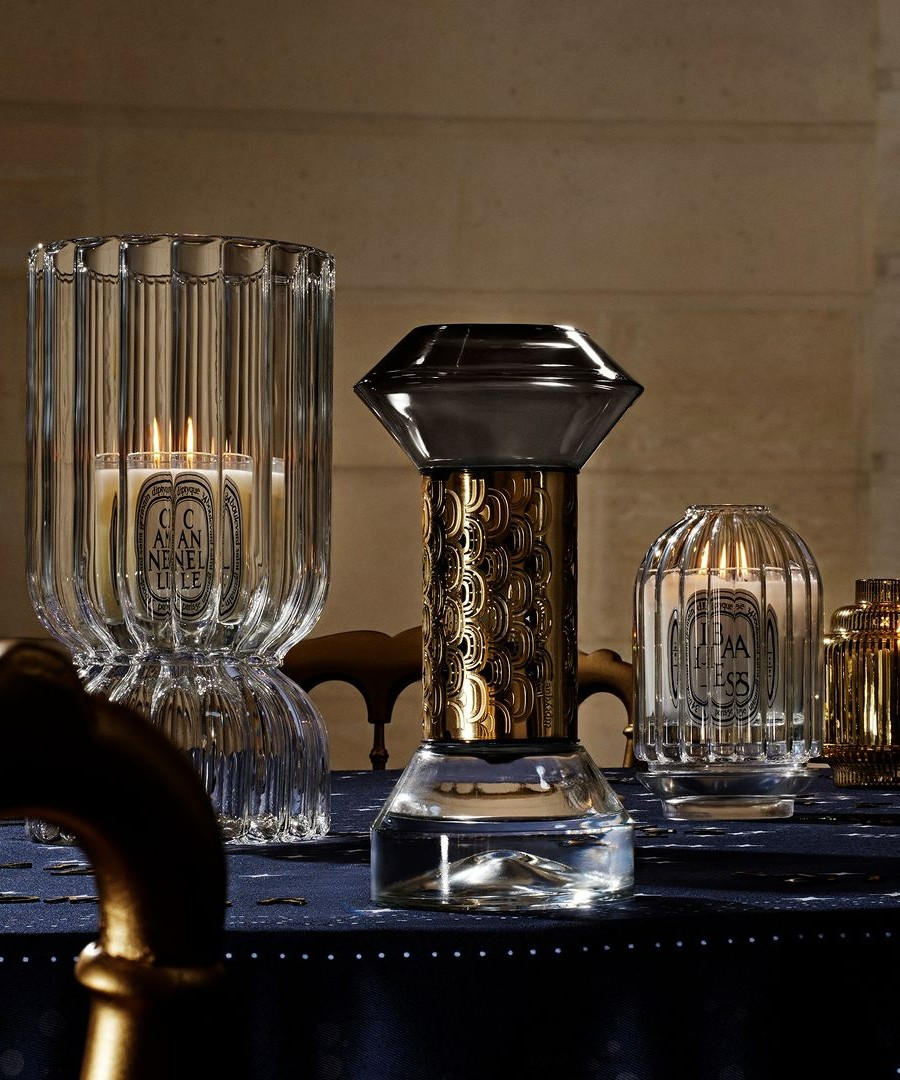 Diptyque candles placed atop a fancy table setting. Crystal vases surround them.