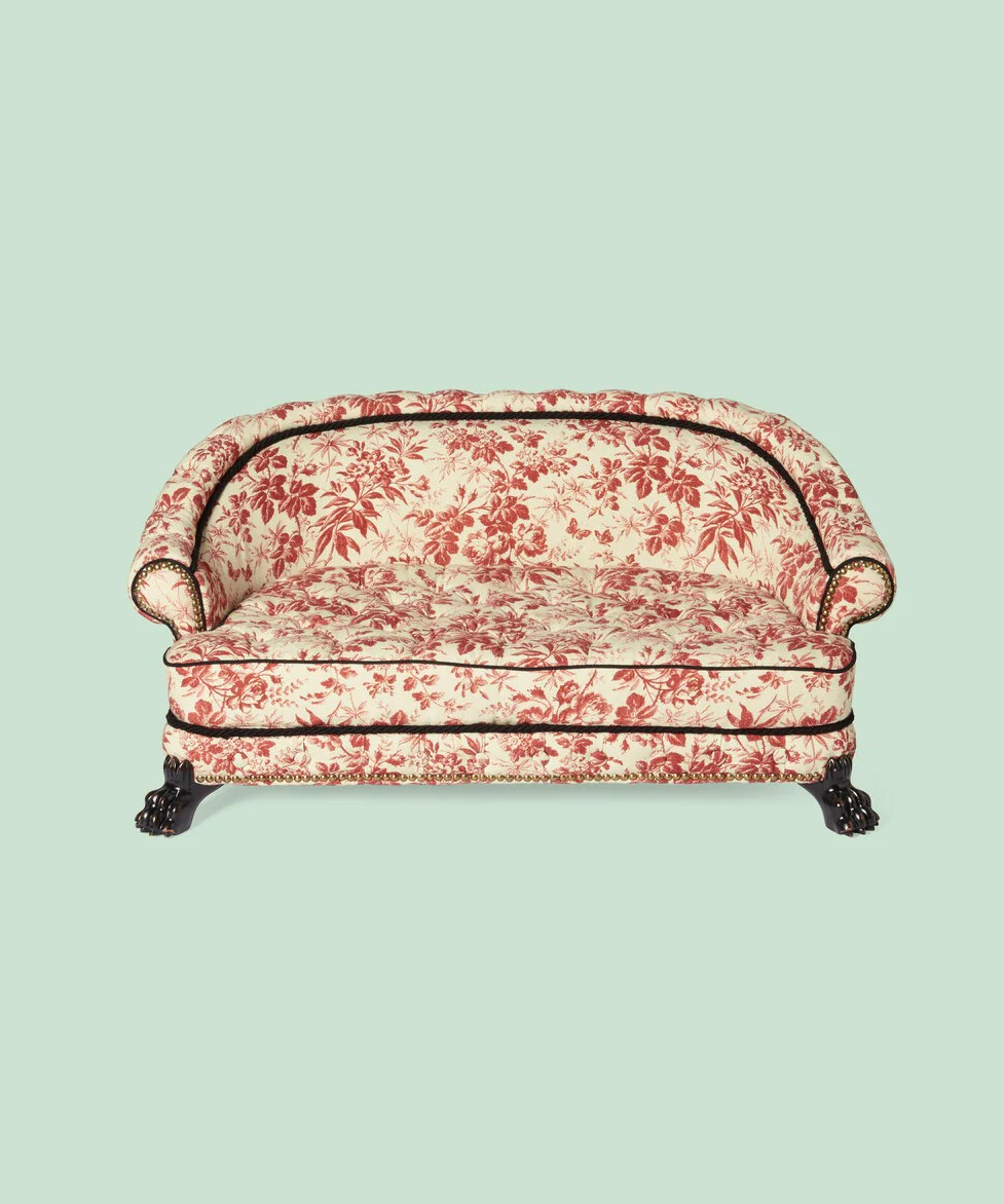 Image of a Gucci dog bed, shaped like a couch. It features an ivory and brick red Herbarium print and features black paws as the base.