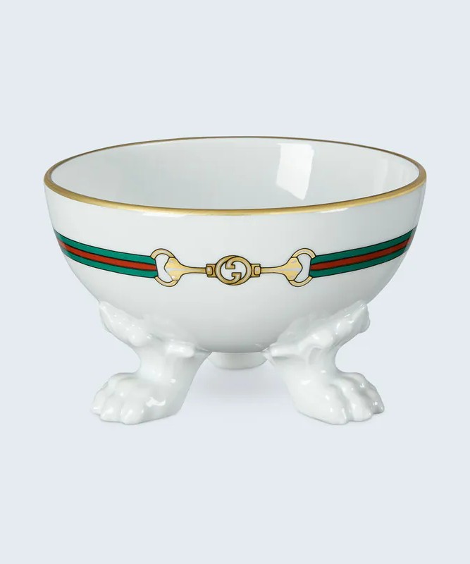 Gucci pet bowl made of white porcelain featuring an interlocking G Horsebit and green and red web print. Three paws sit at the base of the bowl.