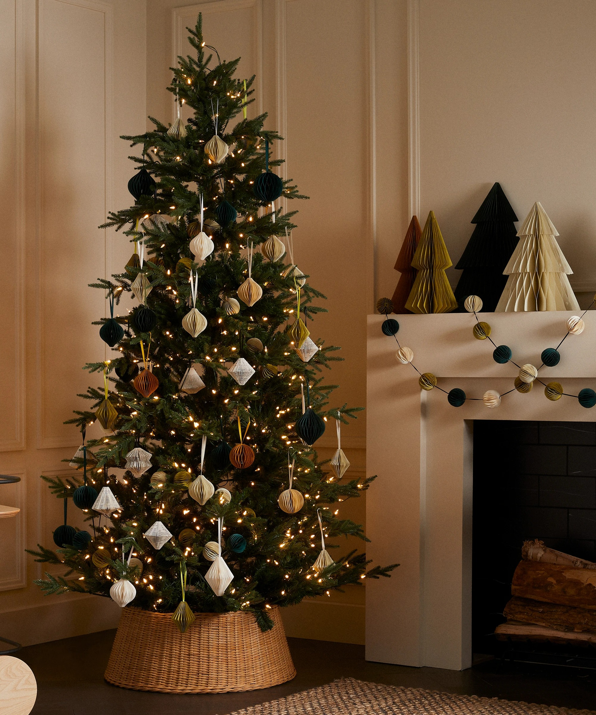 A Christmas tree decorated with a mix of ornaments, ribbons and lights. The tree is displayed in a room with a white wall and it standing beside a white fireplace adorned with paper Christmas trees and paper garland.