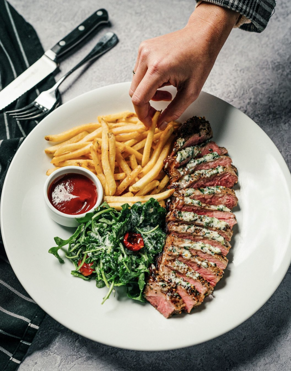 A flat lay image of fries, sliced steak, salad and a small bowl of ketchup. A hand reaches for a fry.