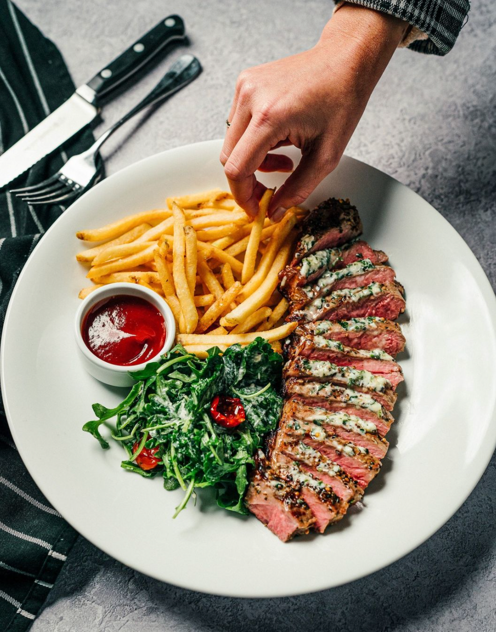 A flat lay image of fries, sliced steak, salad and a small bowl of ketchup. A hand reaches for a fry.