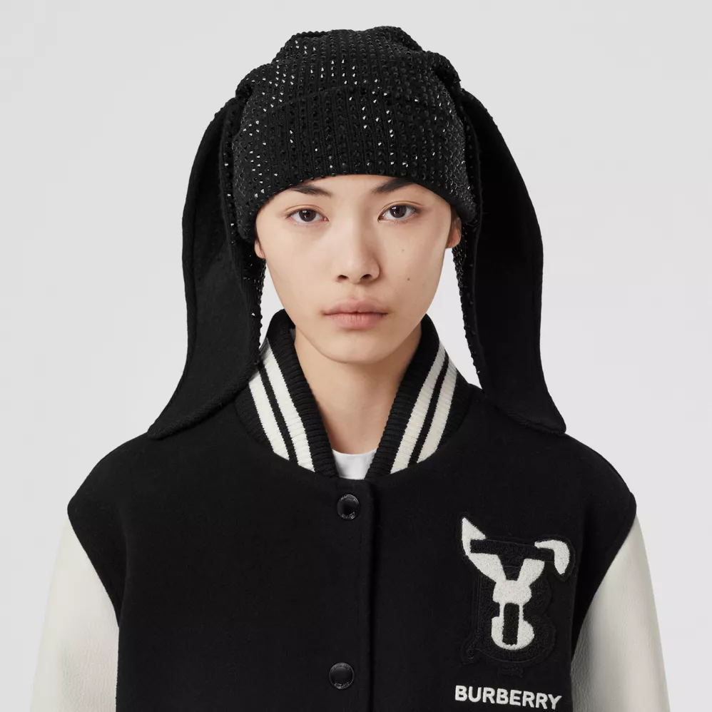 Close-up image of a model wearing a black and white Burberry Lunar New Year varsity jacket with a bunny on the left chest area. The model wears a black toque with long bunny ears.