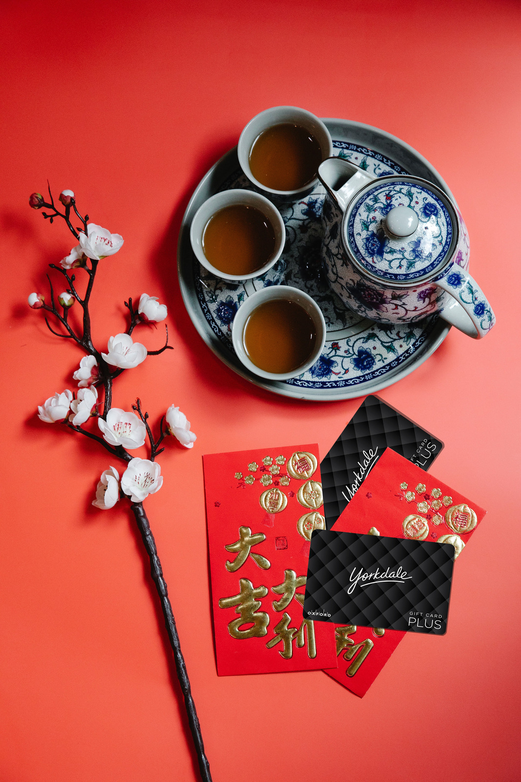 Flat lay image on a red backdrop featuring a tea seat, white florals, lucky red envelopes and a black Yorkdale gift card.