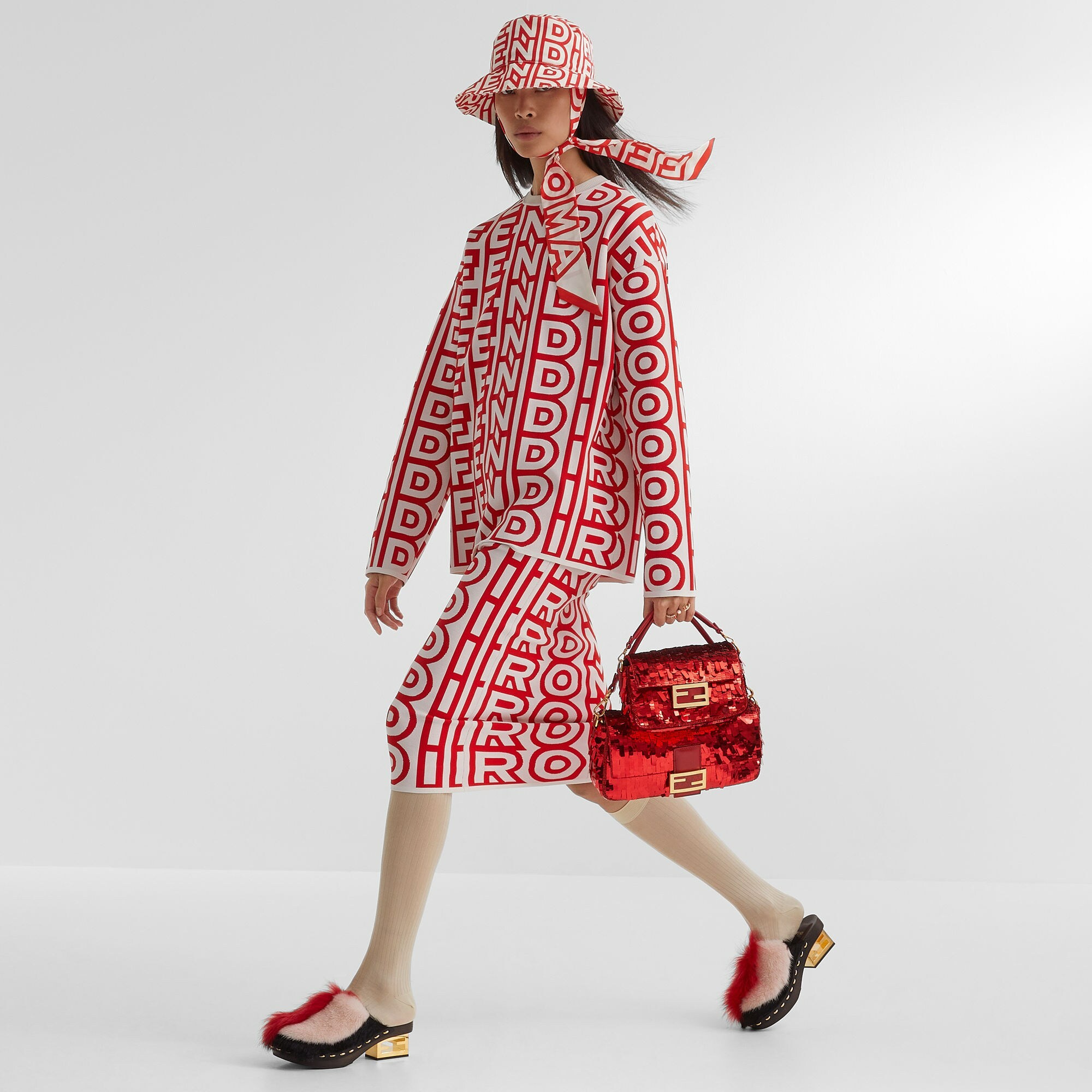 Image of a model wearing a matching red and white fendi set featuring a bucket hat, oversized jacket and skirt. She holds two sparkly red Fendi baguette bags (one is smaller) and wears red, white and black furry clogs.