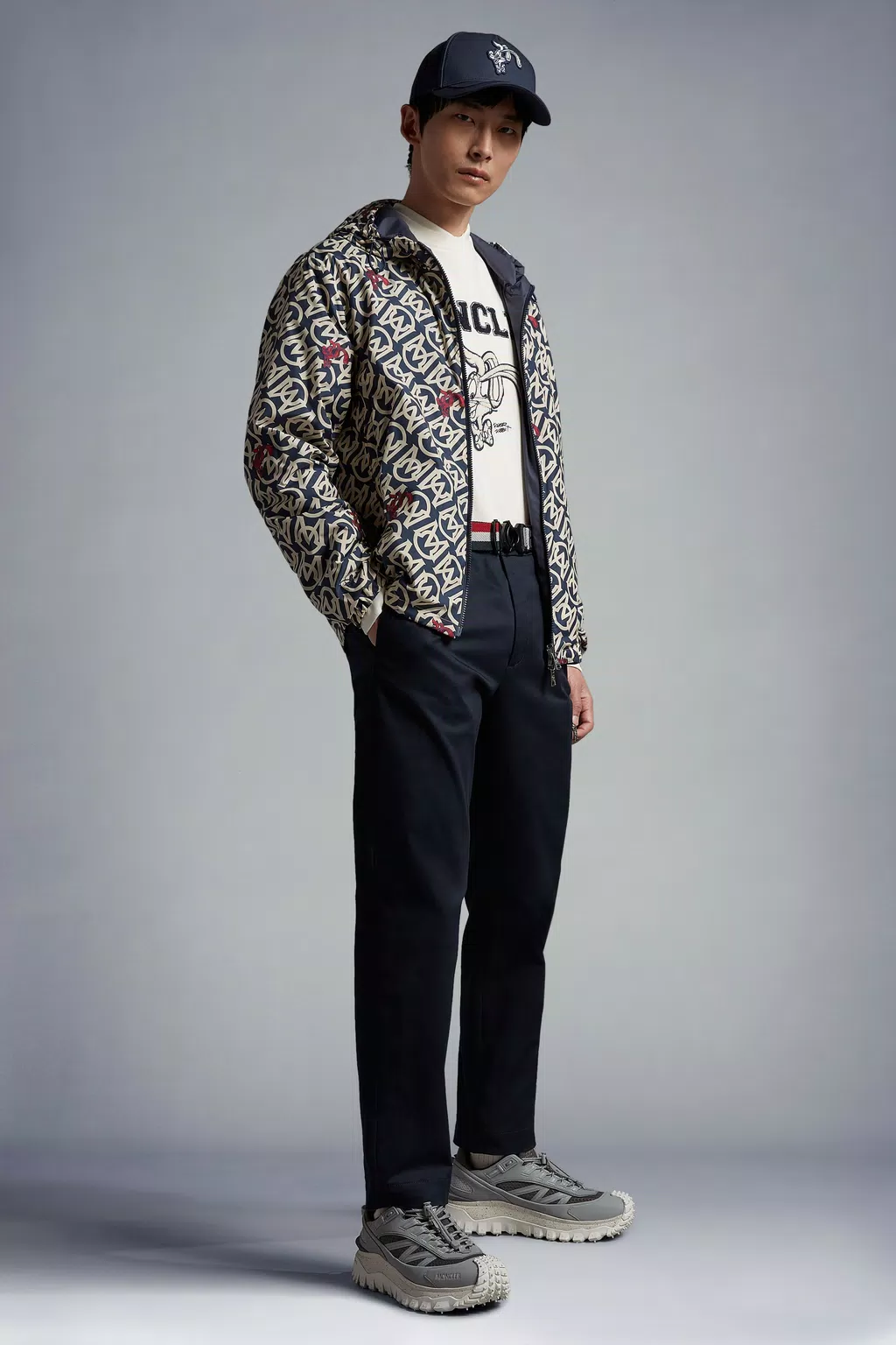 Image of a male model wearing the following Moncler items: a baseball cap with a bunny motif, a white T-shirt with a bunny motif, a printed blue and white zip up sweater with bunnies throughout, navy blue trousers and grey sneakers.