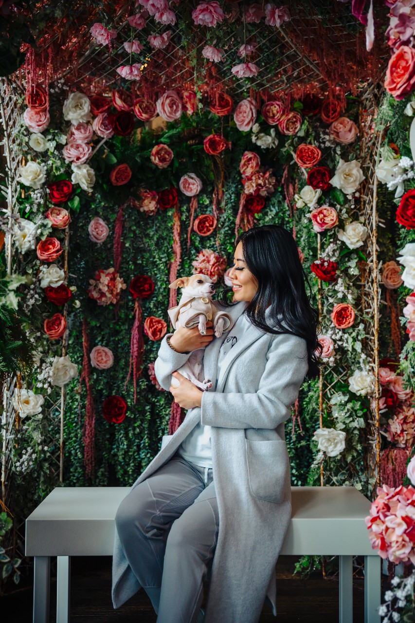 Image of a woman holding a small pet dog wearing a beige button up coat. The woman is sitting on a bench in front of a floral backdrop.