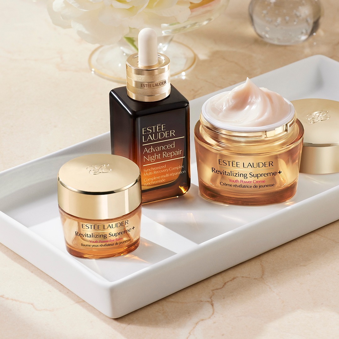 The image is of three skincare products on a white tray. The first product to the left is a small closed, golden jar. The middle product is an ombre brown to golden bottle with a dropper. The final product on the right is a medium-sized open, golden jar with a whipped cream-like product with the lid directly behind it.