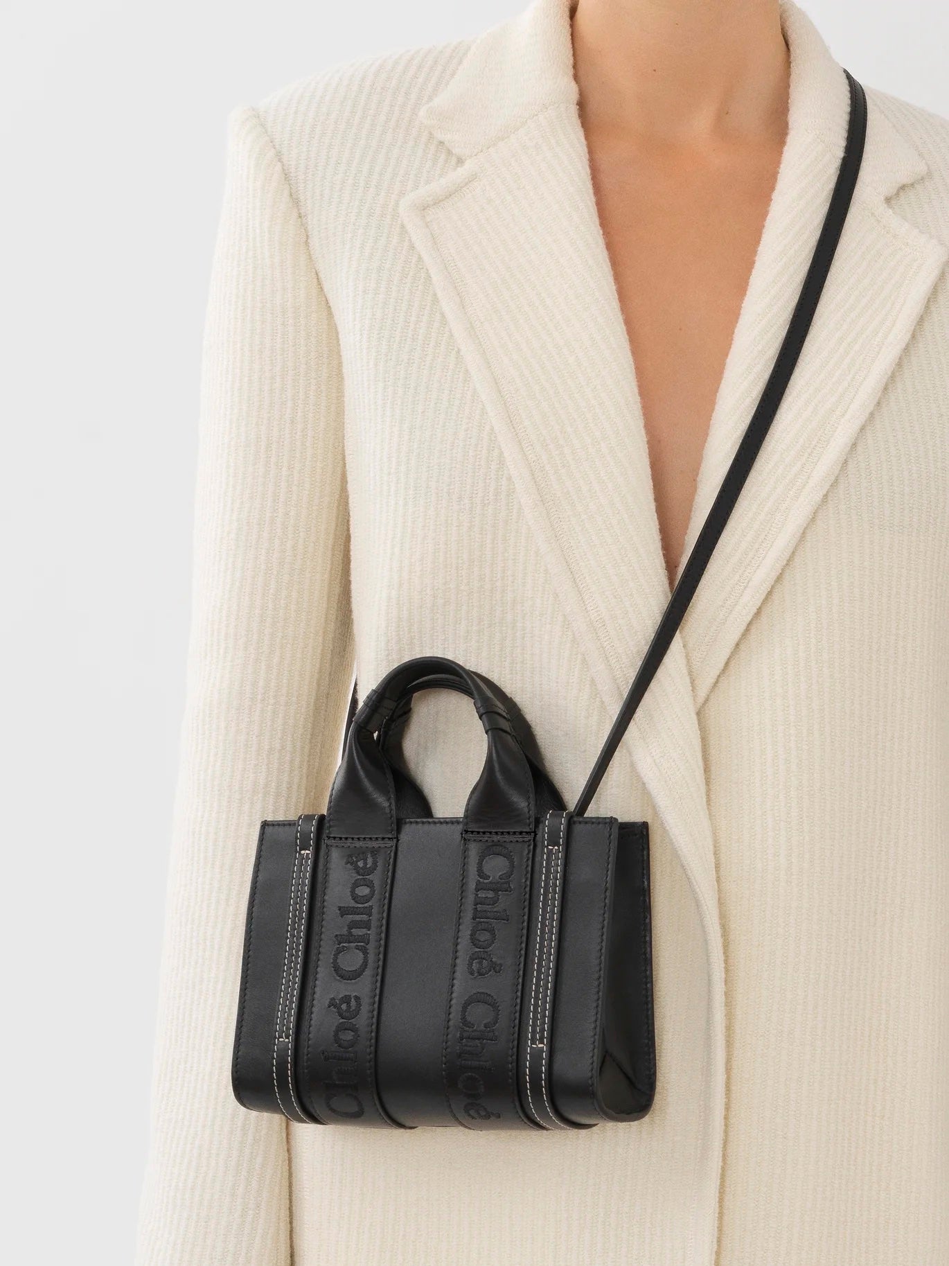 A model wearing a cream wool jacket with a small black purse. The black purse has a long strap sitting over the models right shoulder. The black purse has two small handles on the top with brand detailing.