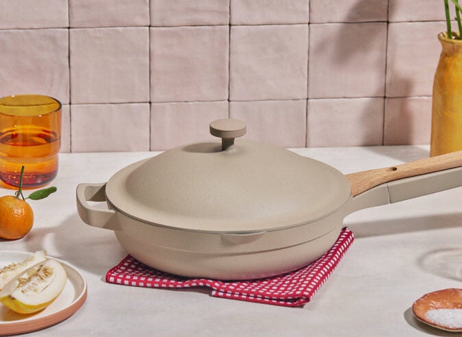 A neutral-coloured pan with a matching lid and wooden handle sitting on a red and white checkered cloth. The background has an assortment of orange, yellow, and white props such as a glass, orange, lemon cut on a plate, and small bowl with salt.