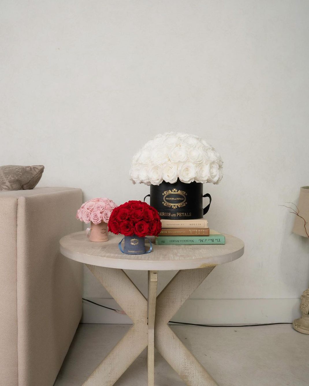 Beside a neutral-coloured couch is a similar colour three-legged table. On top of the table are three coffee table books stacked on top of each other. On the very top of the coffee table books is one of the floral arrangements in a black box with white roses. To the left is a pink box with pink roses, and to the right is a grey box with red roses.
