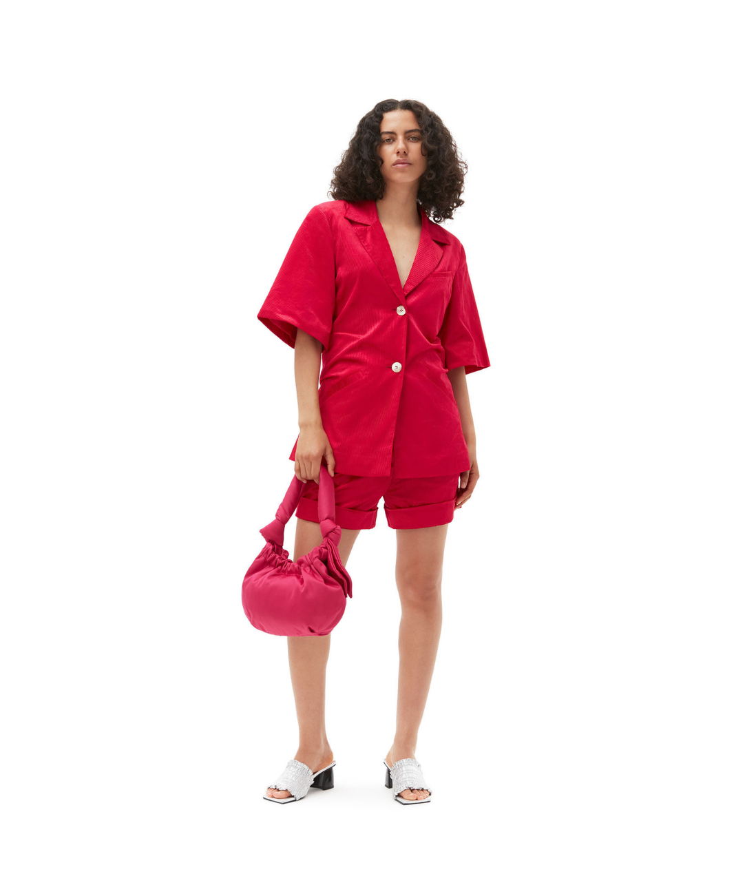 Image of a model wearing a matching red GANNI set featuring an oversized, short sleeve blazer and matching shorts. she holds a red bag and wears white heeled mule sandals.