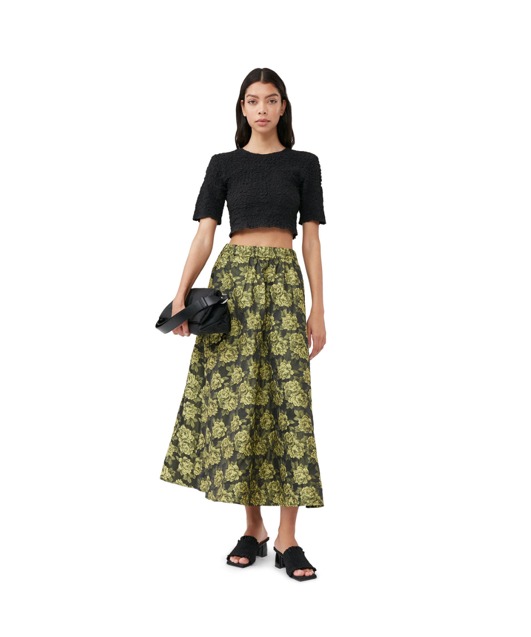 An image of a model on a white backdrop wearing the following GANNI items: a black textured cropped t-shirt, a high-waisted A-line skirt black and yellow skirt with a floral print, a black handbag and black heeled mule sandals.