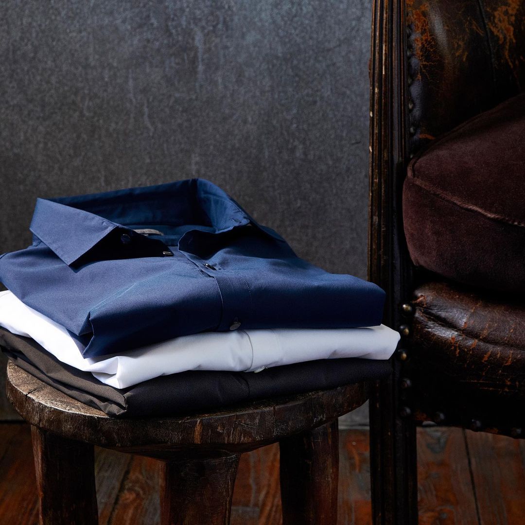 An image of three dress shirts (blue, white and black) folded and stacked neatly onto a wooden bench.