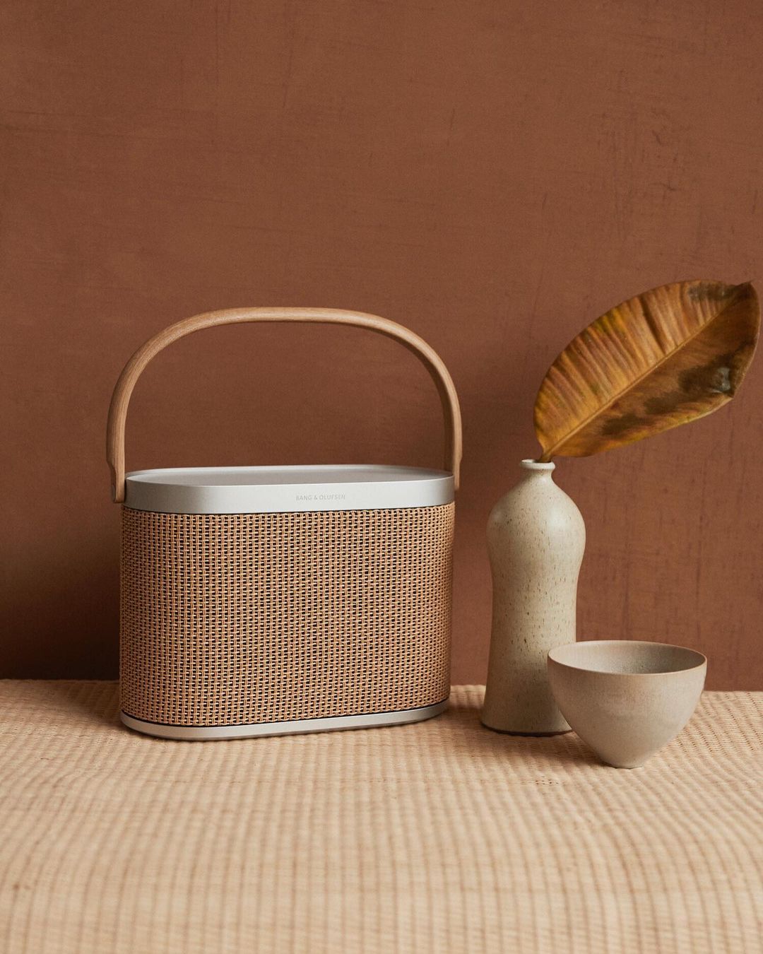 Lifestyle image of a bronze and silver Bang Olufsen speaker with a wooden handle. It sits on table covered in a beige table cloth. Beside it are some ceramics for decoration.