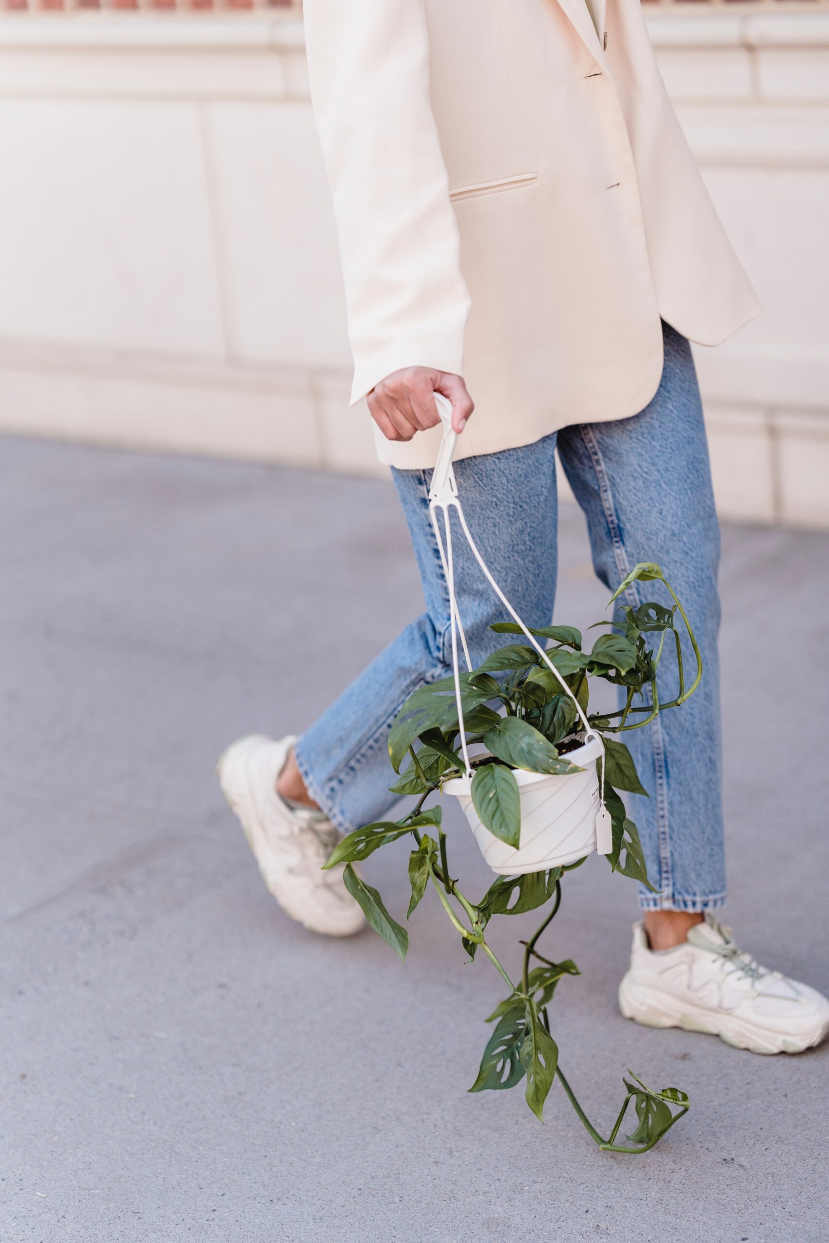 close up image of a woman wearing a white blazer and jeans and white sneakers walking while holding a plant