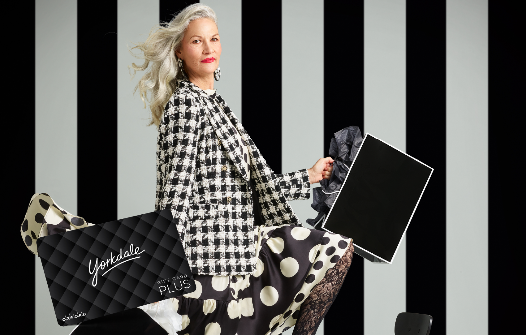 promotional image for a Yorkdale gift card. It shows a woman wearing a black and white houndstooth blazer and black and white polka dot skirt holding black and white shopping bags. A Yorkdale gift card is also in the image