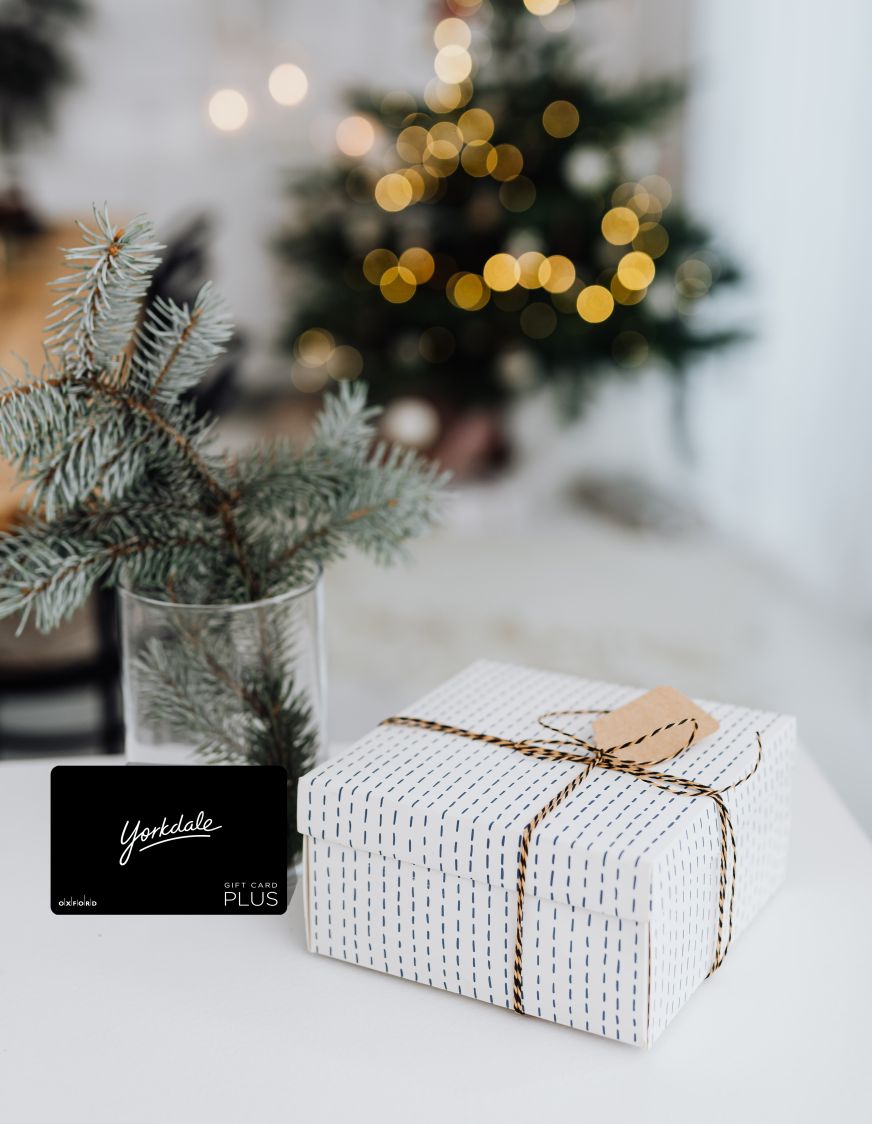 promotional image featuring a black yorkdale gift card. beside the gift card is a present wrapped in white wrapping paper and behind the gift card is a vase with pine tree branches. a christmas tree can be seen in the background