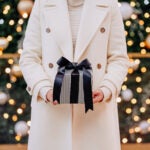 Woman with a white coat, holding a holiday grey gift with a dark bow.