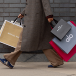 Woman with a textured brown coat holding shopping bags from Michael Kors, Coach and Alo.